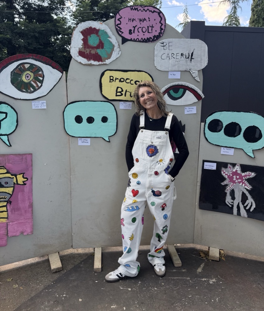 Kara in hand-painted overalls in front of a wall of student cardboard art
